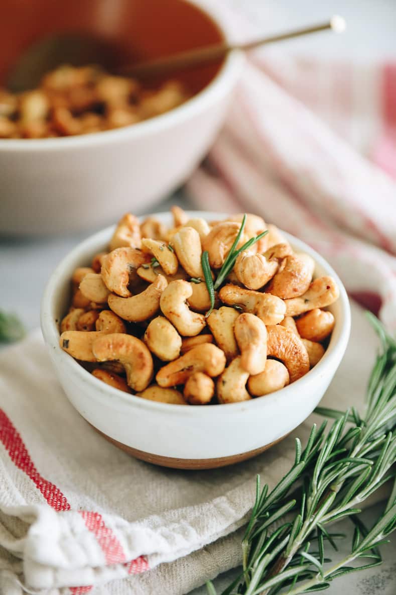 A white bowl of roasted cashews with rosemary.