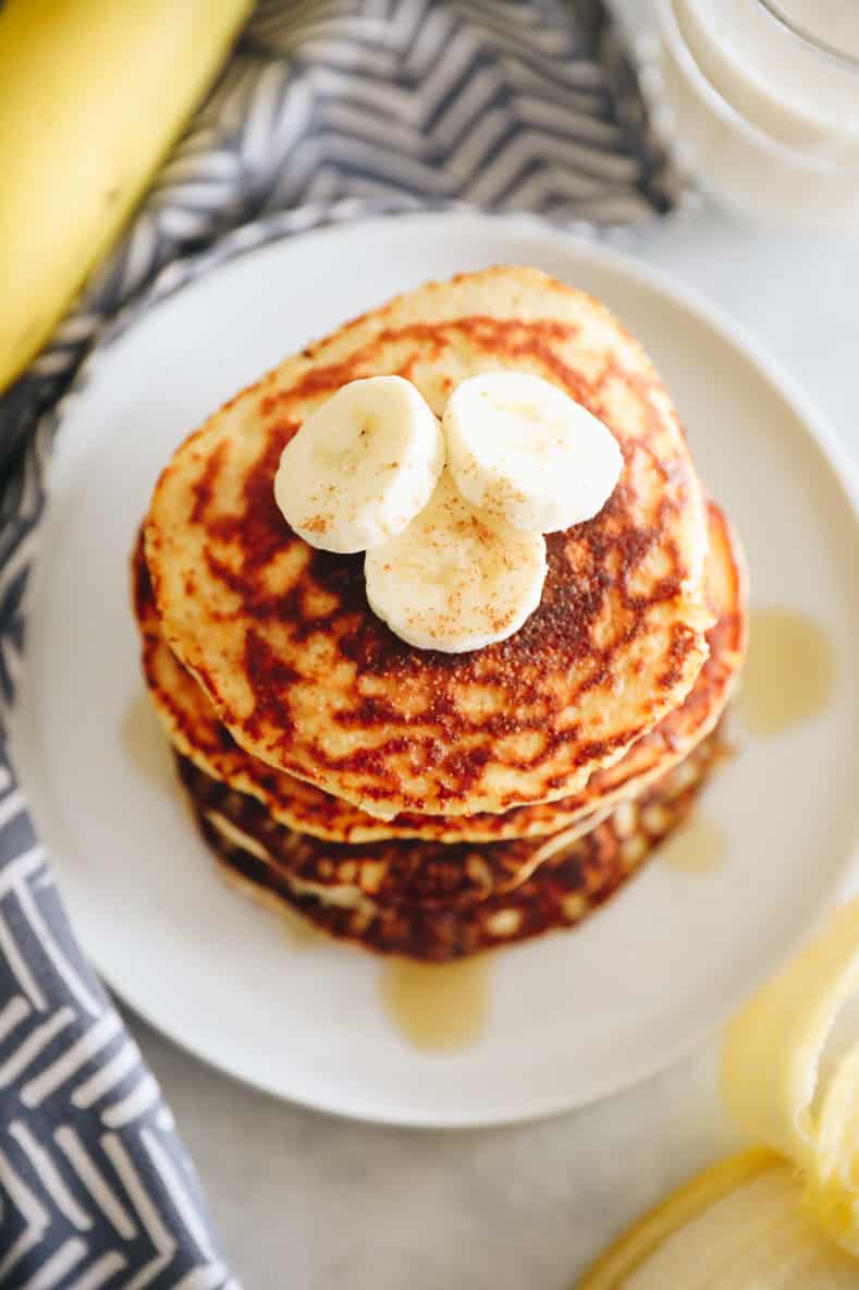 Angled overhead shot of banana protein pancakes on a white plate. Pancakes are drizzled with maple syrup and topped with banana slices.