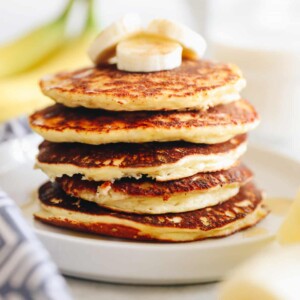 Close up shot of banana protein pancakes stacked on a white plate. Bananas in foreground and background of the image.