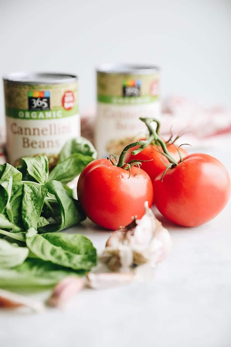 Tomatoes and fresh basil in focus. Garlic cloves and cans of cannellini beans are in the foreground and background.