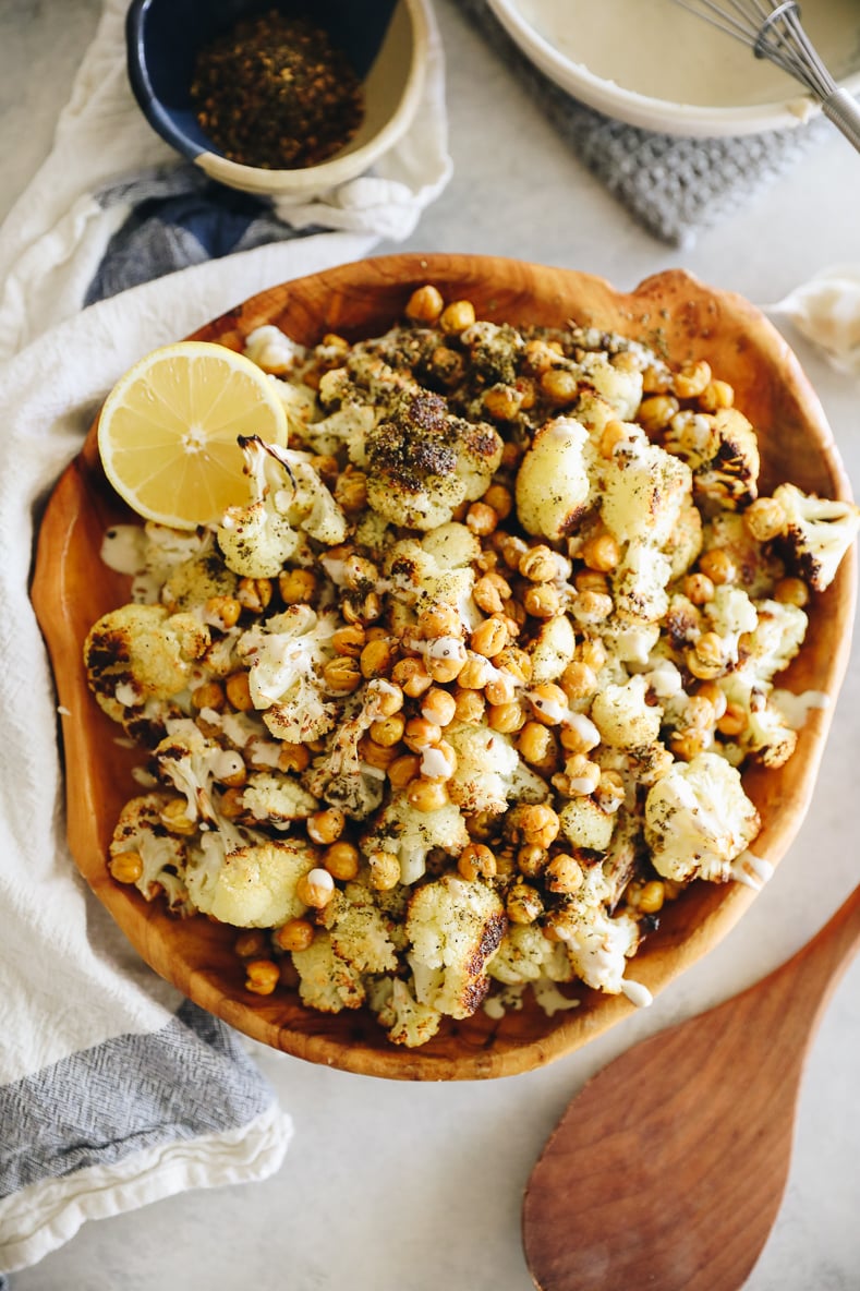 A wooden serving bowl filled with roasted cauliflower and chickpeas. Wooden spoon, za'atar and tahini sauce surround the bowl.
