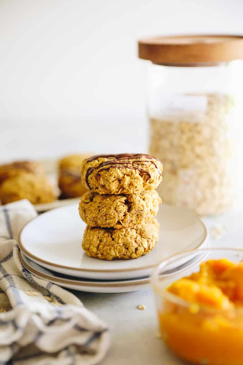 Straight on shot of a stack of pumpkin breakfast cookies on a white plate. Bowl of pumpkin in the foreground and jar of oats in the background.