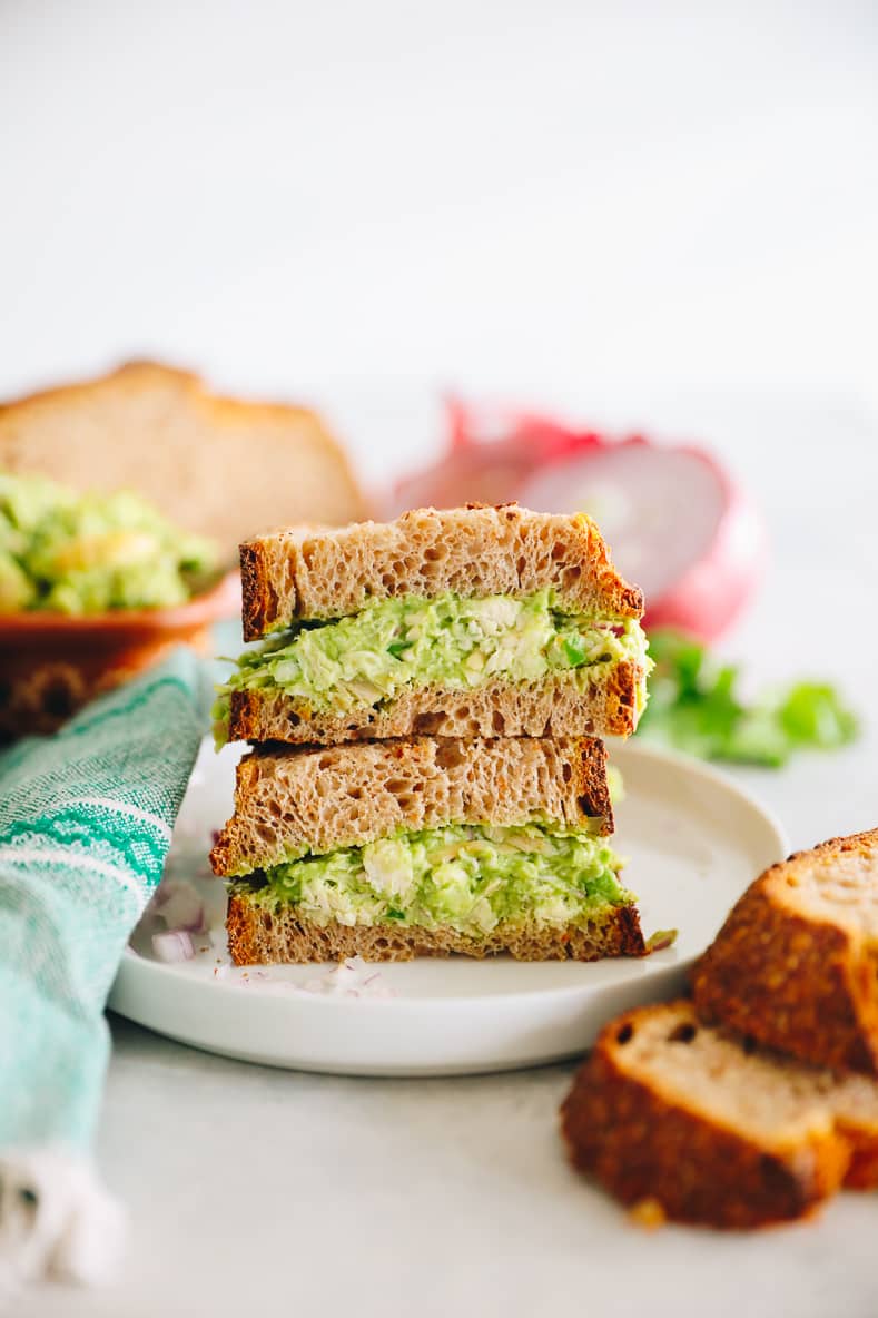 Guacamole chicken salad sandwich on thick slices of wheat bread. Two halves of the sandwich are stacked on top of each other with the middle facing the camera. Ingredients in the foreground and background of the image.