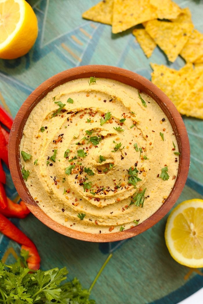 Overhead shot of lemon hemp hummus in a wooden serving bowl surrounded by fresh lemon halves, sliced red bell peppers and tortilla chips.