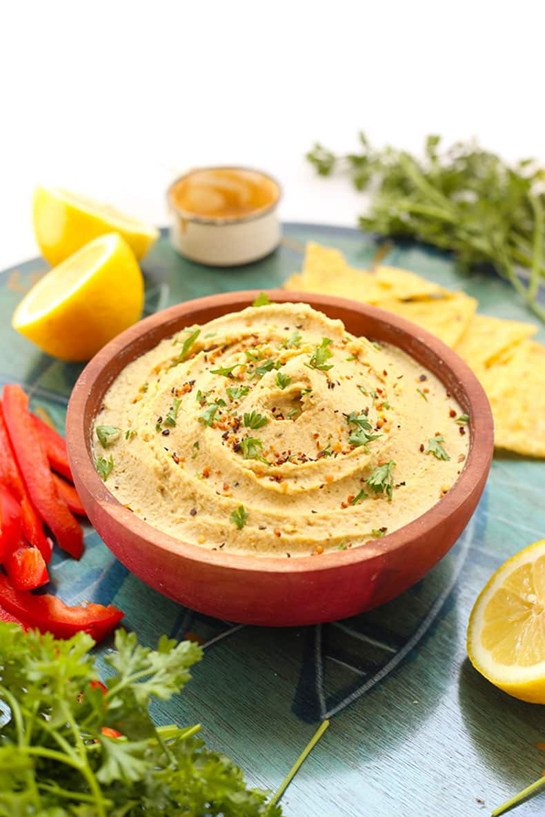 Wooden bowl of lemon hemp hummus surrounded by fresh lemons, chips, herbs and chopped red bell peppers.