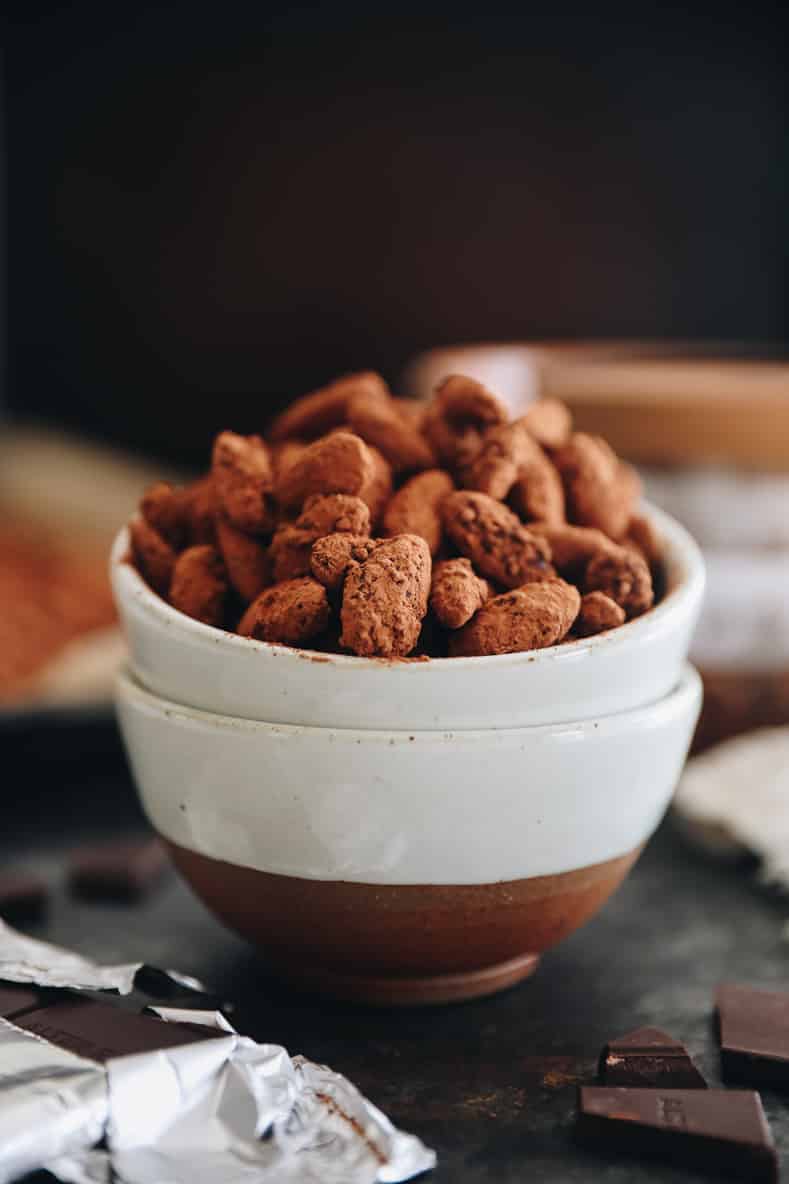 Dark chocolate almonds in a white ceramic bowl. Chocolate pieces in the foreground.