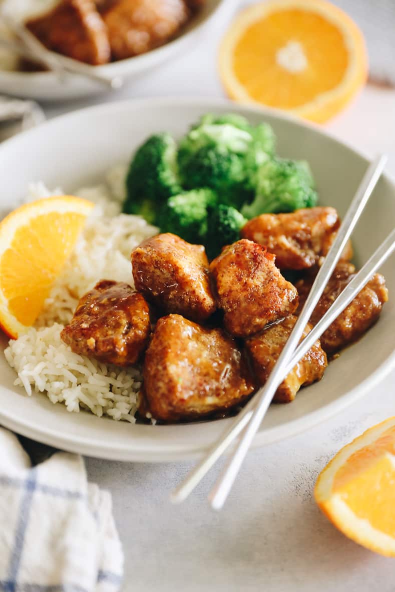 Angled shot of sticky orange chicken on a white plate with rice and broccoli. Silver chopsticks crossed on top.