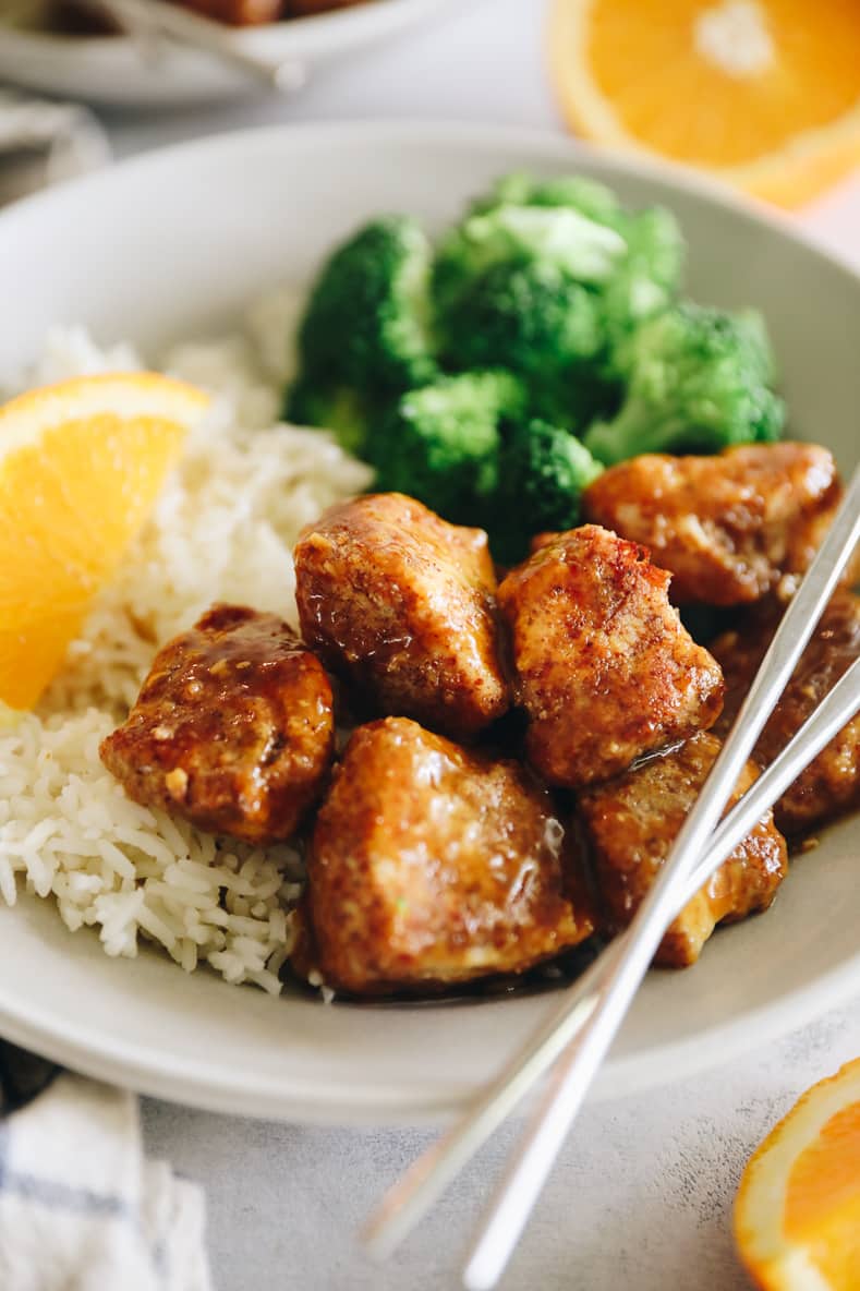 Angled shot of sticky orange chicken on a white plate with rice and broccoli. Silver chopsticks crossed on top.