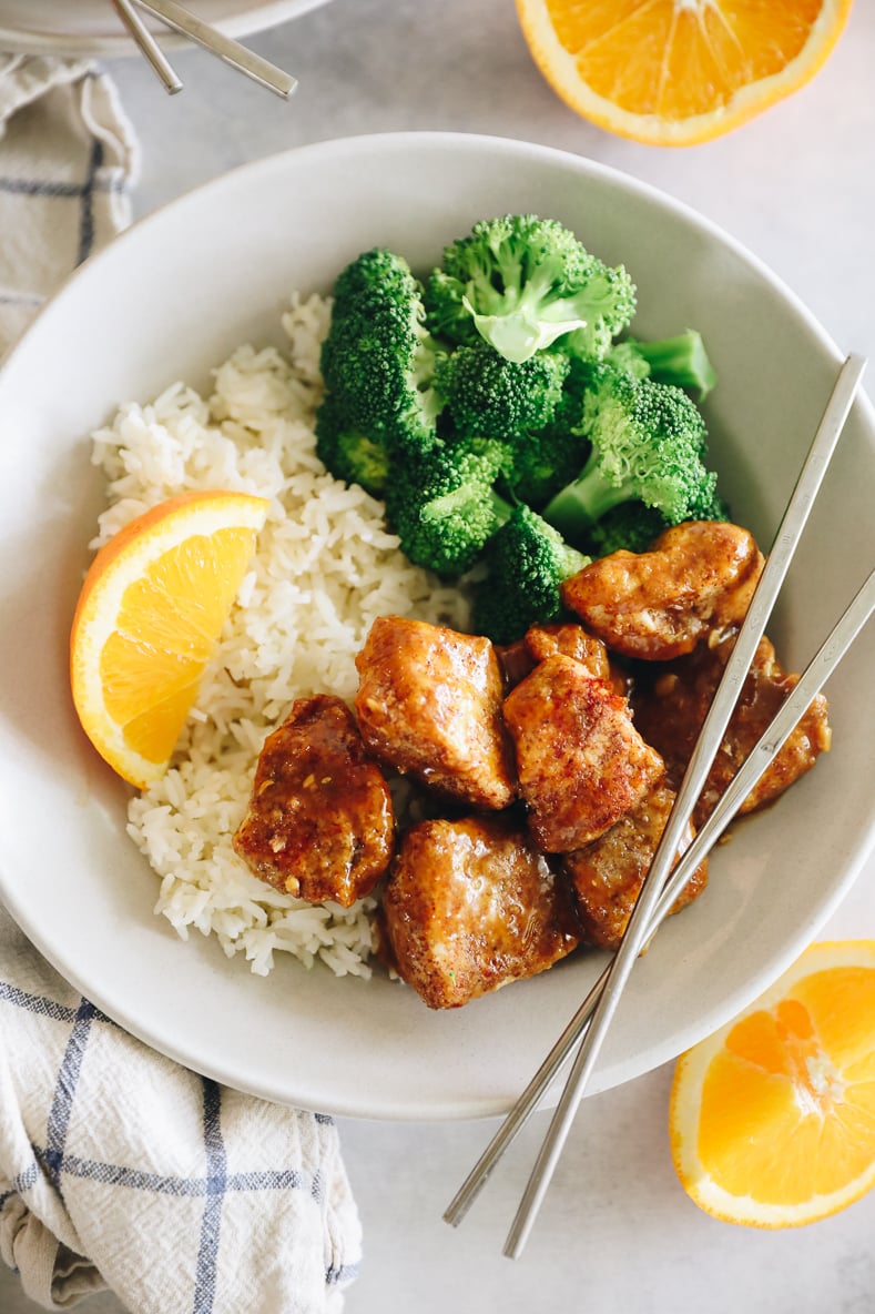Sticky orange chicken on a white plate with rice and broccoli. Silver chopsticks crossed on top.