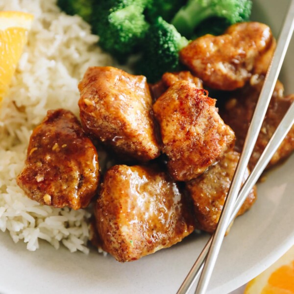 Close up of sticky orange chicken on a white plate with rice and broccoli. Silver chopsticks crossed on top.