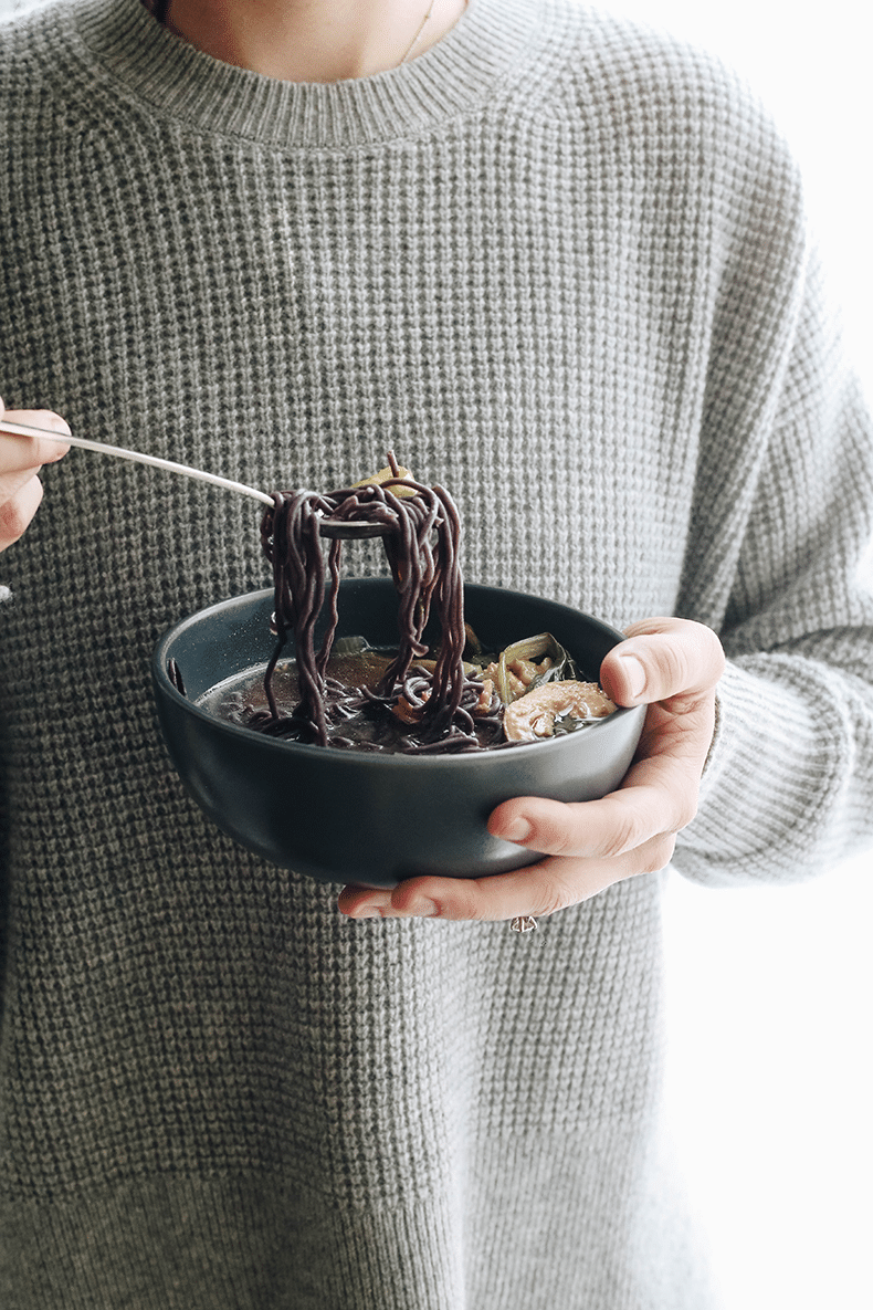 Person in a grey waffle sweater holding a bowl of healing soup. Silver spoon taking a scoop of noodles and vegetables out of the soup.