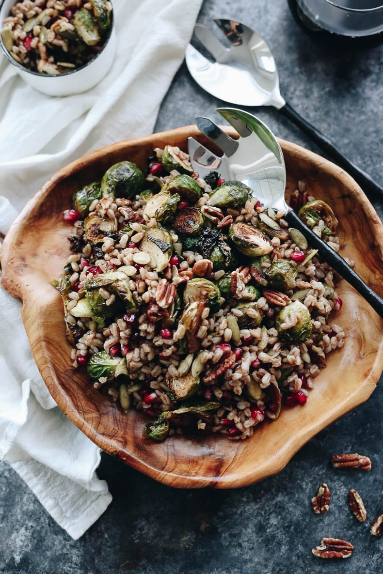 roasted brussel sprouts and farro salad in wooden bowl topped iwth pecans, pomegranate seeds and pumpkin seeds.