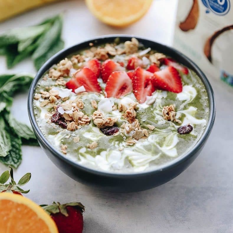 Matcha mint smoothie bowl topped with granola, yogurt and fresh berries. Various ingredients blurred in the background. 