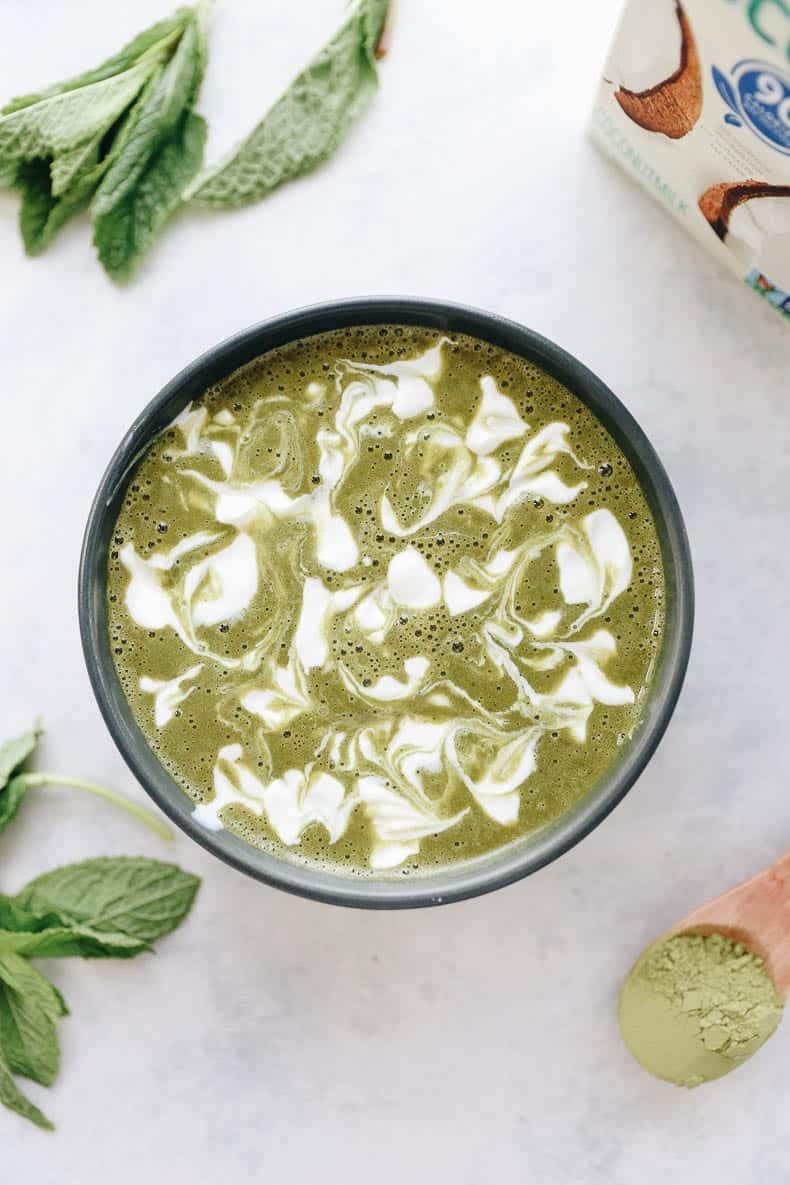 Overhead shot of a matcha mint smoothie bowl swirled with coconut milk. Mint, matcha and coconut milk surround the bowl.