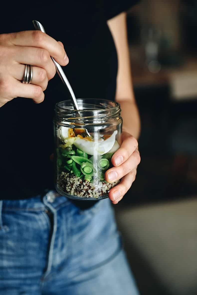 Hand holding jar filled with quinoa, snap peas and eggs. Silver utensil coming out of jar. 