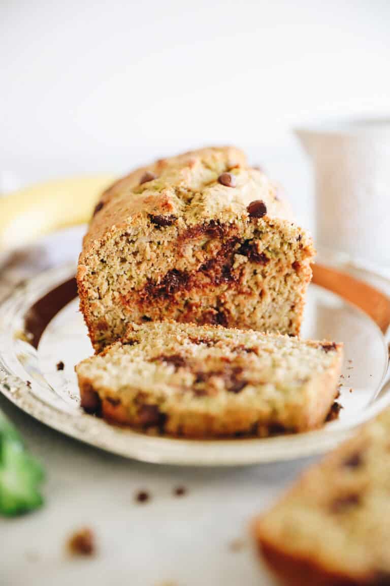 Loaf of zucchini banana bread sitting on a silver platter. One slice of bread laying in front of loaf.