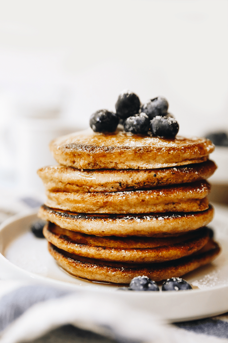 Stack of whole wheat pancakes on a white plate topped with fresh blueberries and a sprinkle of powdered sugar. 