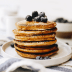 Stack of whole wheat pancakes on a white plate topped with fresh blueberries and a sprinkle of powdered sugar.