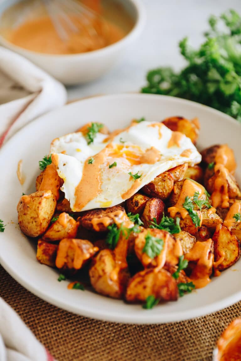 Plate of patatas bravas topped with a fried egg, a drizzle of sauce and fresh herbs.