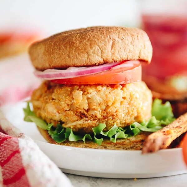 Quinoa veggie burger topped with lettuce, tomato, onion on a whole wheat bun. Burger is on a white plate and surrounded by french fries.