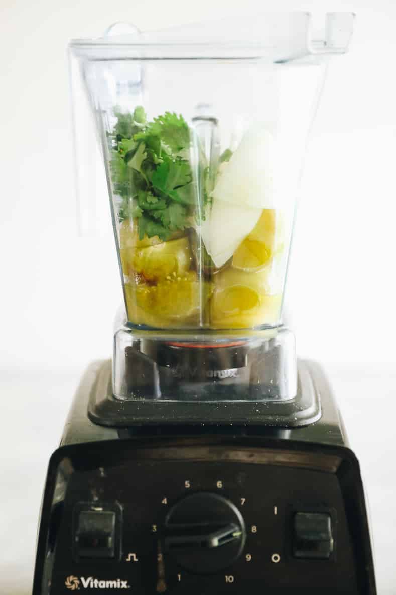 Straight on shot of a Vitamix blender filled with green salsa ingredients.