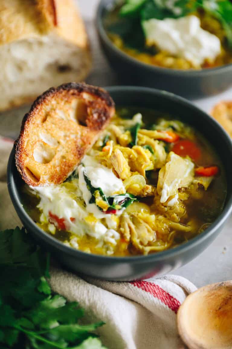 Angled shot of chicken lentil soup in a dark grey bowl. Soup is topped with sour cream and a piece of crunchy bread.