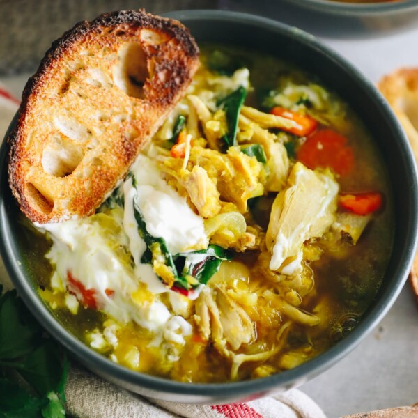 Close up of chicken lentil soup in a dark grey bowl. Soup is topped with sour cream and a piece of crunchy bread.