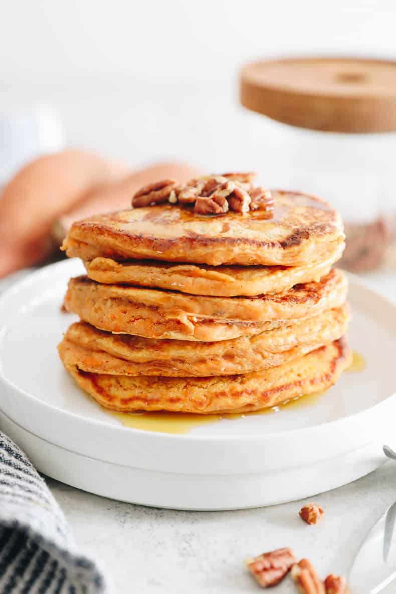 Direct shot of stacked sweet potato pancakes on a white plate.  Topped with crushed pecans and maple syrup.