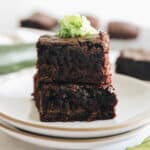 zucchini brownies stacked on a white plate with grated zucchini on top.