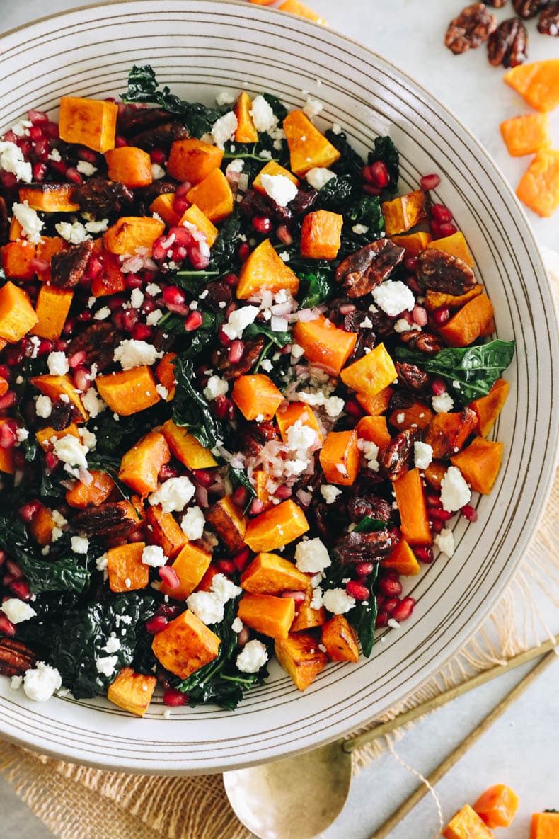 Upclose overhead image of a kale fall salad topped with roasted butternut squash, pomegranate seeds, candied pecans and feta cheese.