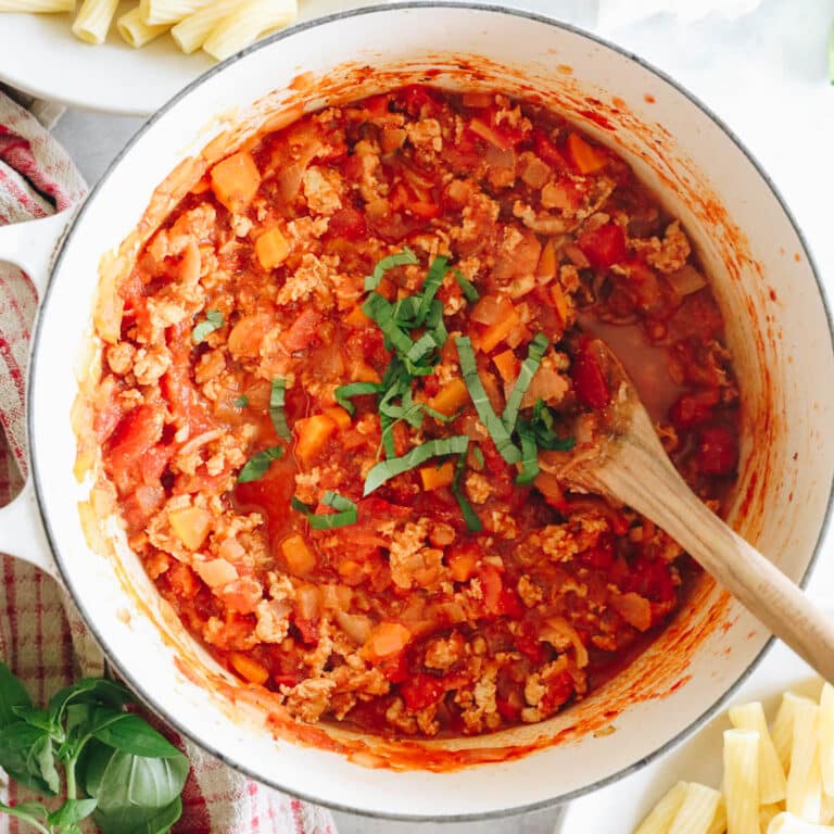 overhead image of turkey bolognese in a white dutch oven sprinkled with basil and a wooden spoon.