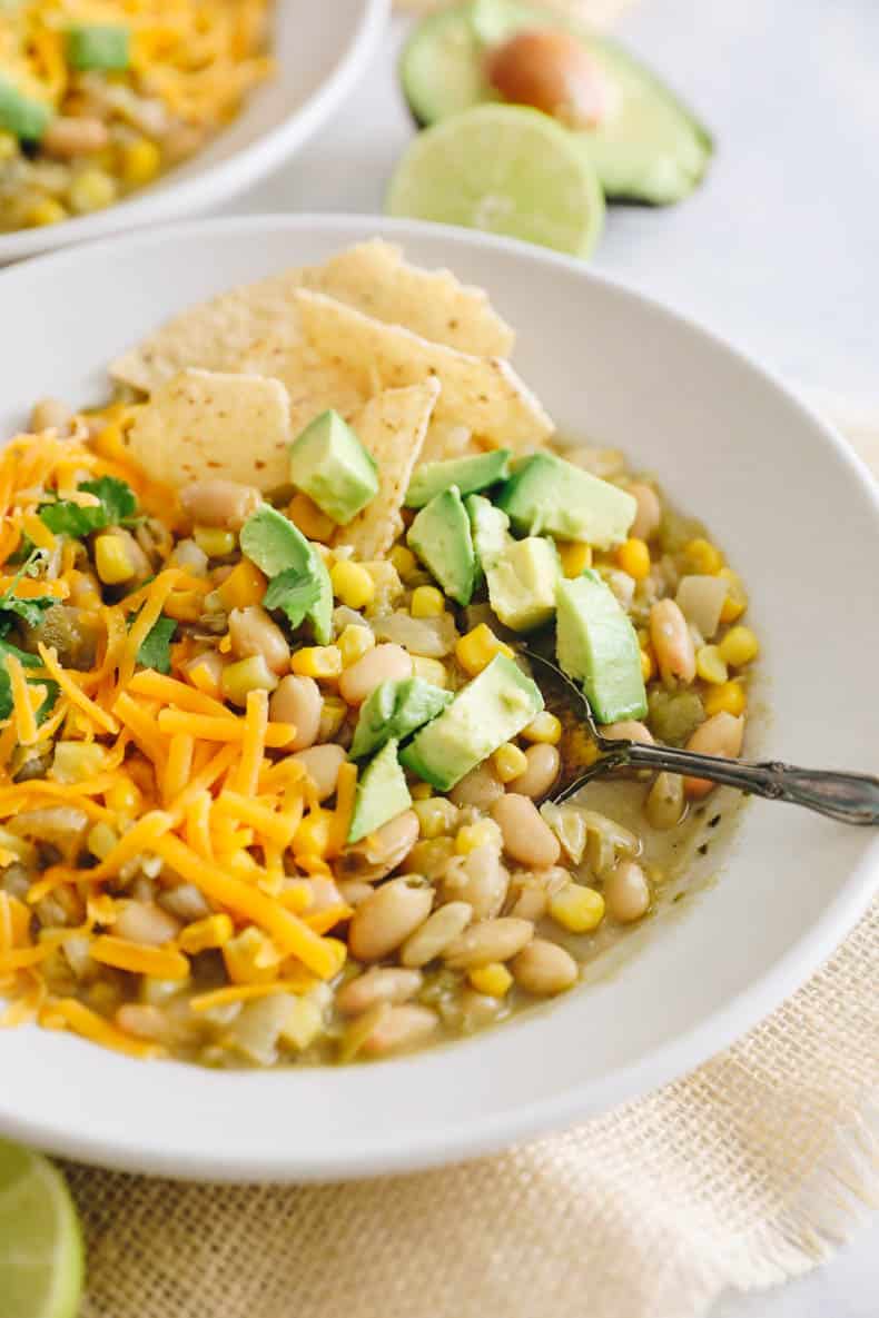 white bean chili topped with avocados, shredded cheese, cilantro and tortilla chips in a white bowl.