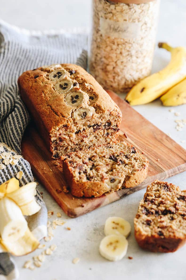 oat flour banana bread on a cutting board with chocolate chips and sliced bananas.