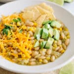 white bean chili topped with avocado, shredded cheese and tortilla chips in a large white bowl.