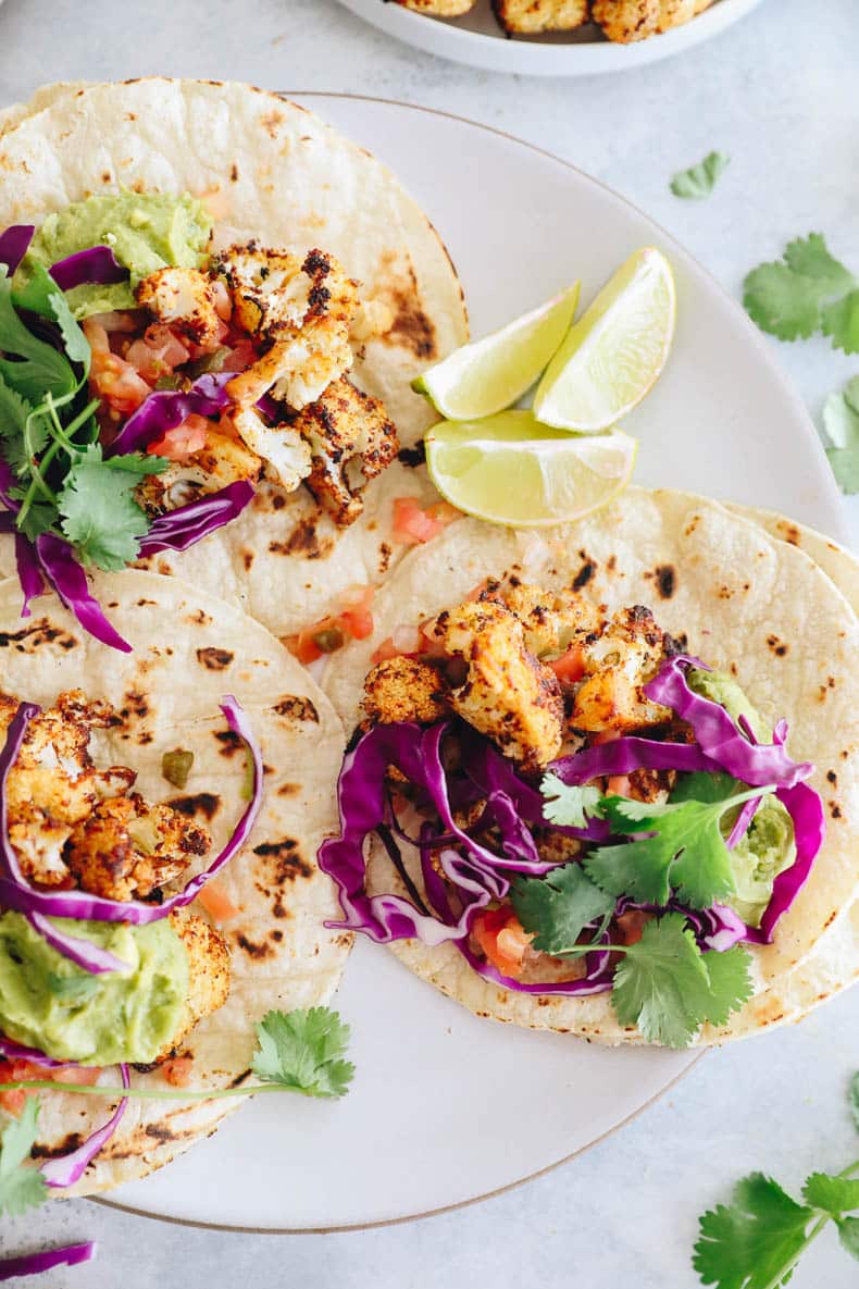 3 cauliflower tacos on a plate with lime slices, pico de gallo, guacamole and red cabbage.